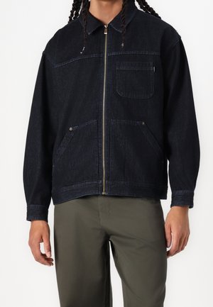 Man wearing black zip-up denim jacket with collar and three front pockets, paired with olive green pants, facing forward.