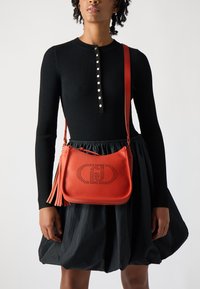 Red leather crossbody bag with perforated logo detail, featuring a detachable fringed accent, paired with a black ribbed long-sleeve top.