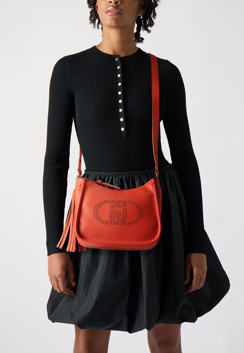 Red leather crossbody bag with perforated logo detail, featuring a detachable fringed accent, paired with a black ribbed long-sleeve top.