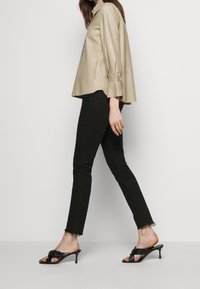 Kvinna som bär en beige långärmad skjorta, svarta skinny jeans med fransar och svarta klacksandaler, som går mot en enfärgad ljus bakgrund.
