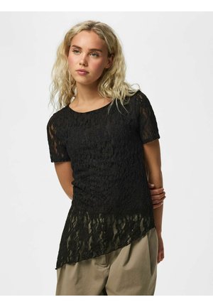 Jeune femme aux cheveux blonds bouclés portant un haut en dentelle noire à manches courtes et un pantalon beige, debout devant un fond blanc.