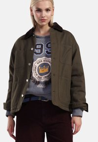 Olivengrøn jakke med et corduroy-krave, store frontlommer og sølvknapper, lagdelt over en grå sweatshirt med grafiske designs.
