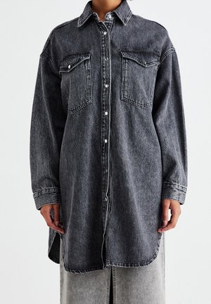 Personne portant une longue chemise en denim gris foncé avec des boutons à l’avant et deux poches poitrine, debout devant un fond uni.