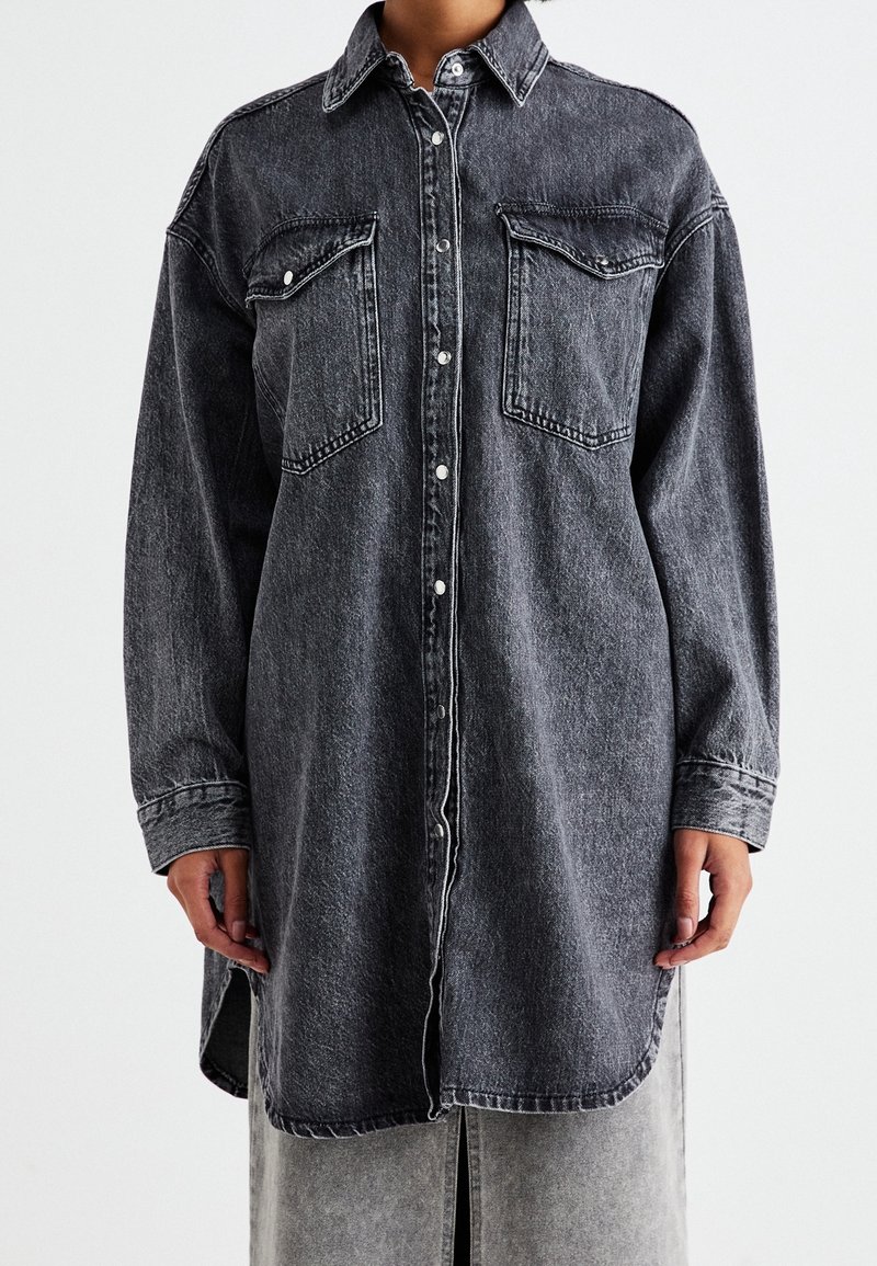 Personne portant une longue chemise en denim gris foncé avec des boutons à l’avant et deux poches poitrine, debout devant un fond uni.
