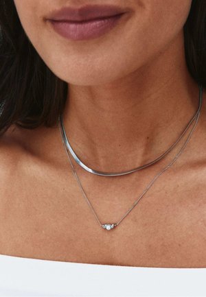 Hals en onderkant van het gezicht van een vrouw die twee zilveren kettingen draagt, één met een klein rond parelhangertje, tegen een wit off-shoulder topje.