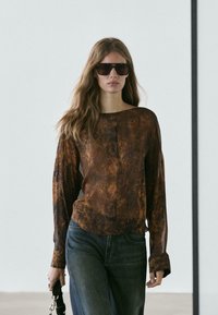 Brun mønstret bluse med lange ærmer lavet af transparent stof, kombineret med blå denimjeans. Modellen holder en sort stroplomme og bærer mørke solbriller.