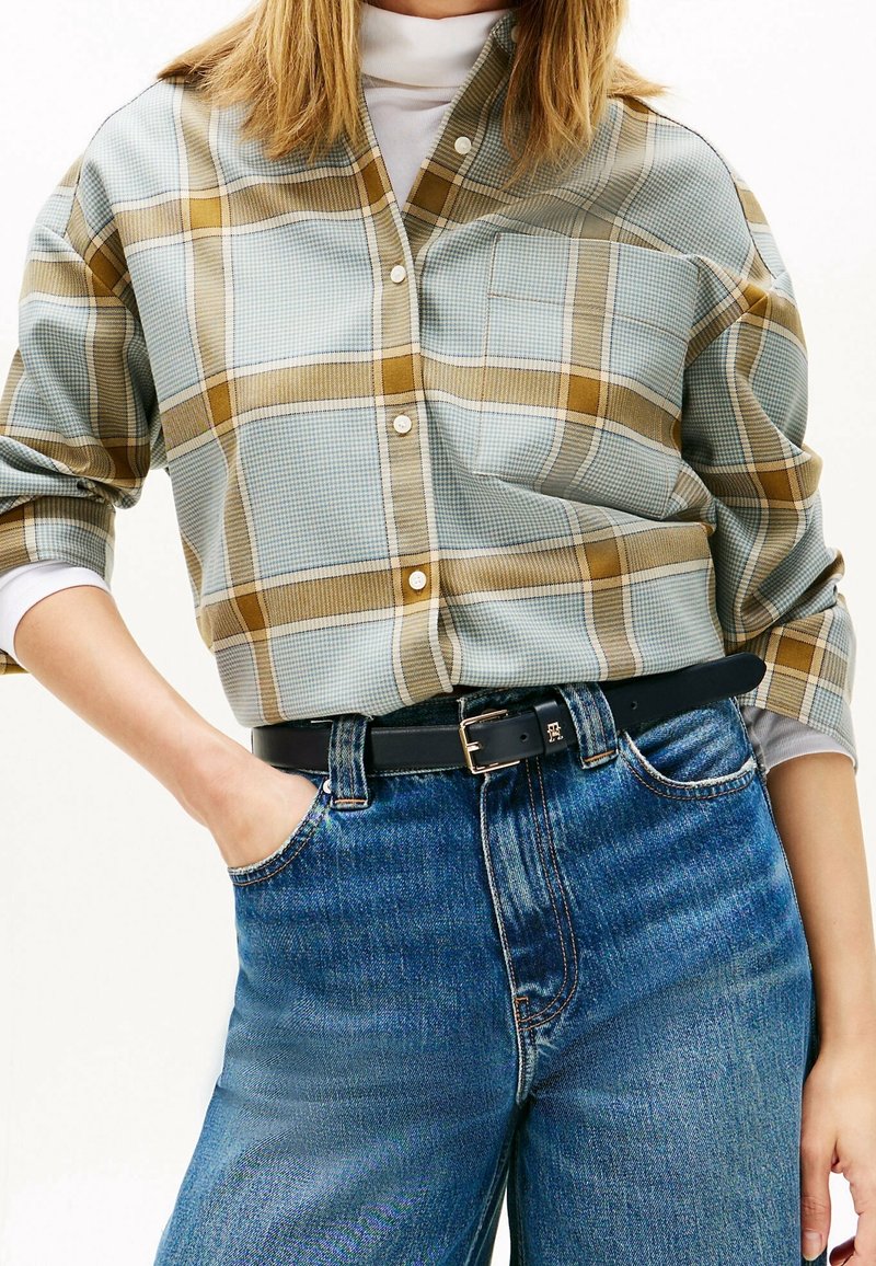 Femme portant une chemise à carreaux beige et bleue boutonnée sur un col roulé blanc, rentrée dans un jean bleu taille haute avec une ceinture noire.