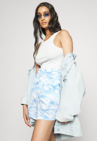 Camisola branca de alças, jaqueta de ganga azul clara e calções estampados com redemoinhos abstratos azuis. Texturas suaves e ajuste relaxado.