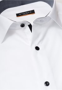 Camicia bianca slim-fit con bottoni neri, un interno del colletto scuro a contrasto, texture liscia e un design di colletto curato.