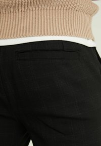 Detailní záběr na osobu v černých texturovaných kalhotách s zadní kapsou a béžovém pleteném svetru přes bílou košili.