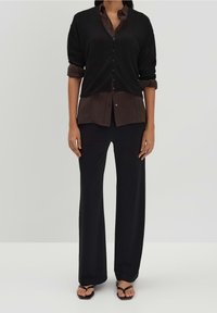 Černá žebrovaná košile na knoflíky přes tmavě hnědou košili s límcem, spárována s černými kalhotami s širokými nohavicemi a černými žabkami.