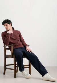 Uomo in camicia a maniche lunghe bordeaux e pantaloni navy seduto su una sedia di legno, reclinato all'indietro con una gamba estesa, indossando sneakers bianche.