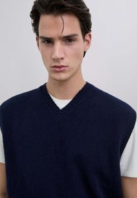 Chaleco de punto sin mangas en color azul marino con diseño de escote en V, hecho de material suave. Llevado sobre una camisa blanca, con bordes acanalados.