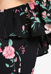 Blusa negra con estampado floral que cuenta con un cuello con volantes, patrones florales vibrantes en rosa y verde, y un discreto detalle de cremallera en el lateral.