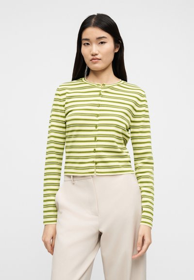 Γυναίκα με μακριά σκούρα μαλλιά που φοράει πράσινη και λευκή ριγέ ζακέτα με κουμπιά και ανοιχτό μπεζ παντελόνι με ψηλή μέση, στέκεται μπροστά σε απλό φόντο.