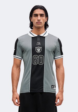 Homme aux cheveux noirs mi-longs porte un maillot de sport Raiders noir et blanc à rayures verticales avec le numéro 60 sur le devant sur un fond uni.