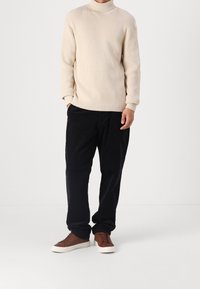Pull à col roulé beige côtelé, pantalon noir ample, et baskets montantes marron à lacets avec semelles blanches sur un fond neutre.