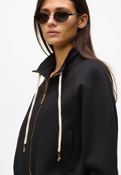 Sweat à capuche noir zippé avec un col montant, des cordons crème ornés d'accents dorés et des poches latérales. Le mannequin porte des lunettes de soleil rondes foncées.