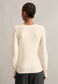 Hellbeige Langarmoberteil mit geripptem Muster und rundem Ausschnitt; taillierte Form, kombiniert mit dunkelgrauen, fließenden Hosen.