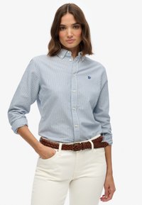Blauw-wit gestreepte overhemden, lichte stof, lange mouwen, kraag. Draagt met een gevlochten bruine riem en crèmekleurige broek.