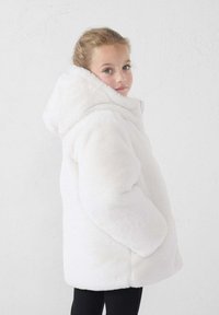 Chaqueta acolchada de piel sintética blanca con una capucha amplia, cremallera frontal y bolsillos laterales. Textura suave y diseño oversize. Leggings negros debajo.