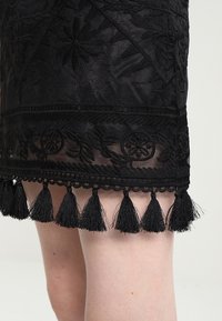 Černá vyšívaná sukně s propracovanými květinovými vzory, průhlednou tkaninou a okrajem z texturovaných černých třásní na lemování.