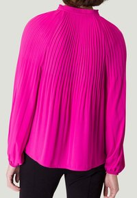 Blouse plissée fuchsia avec un col montant, des manches longues et des poignets élastiques. Tissu lisse avec des plis verticaux dans le dos.