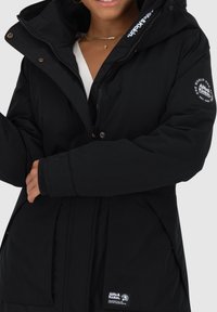 Černá zateplená parka se zipem vpředu, kapucí a žebrovanými manžetami. Má logo nášivku na rukávu a boční kapsy. Měkká textura.