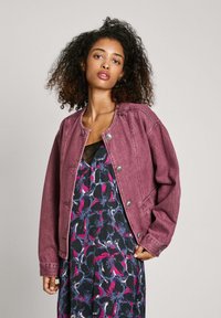 Denim jakna v berry barvi z okroglim izrezom, gumbnim zapiranjem in oversize krojem. Nošena preko obleke s cvetličnim potiskom, ki vključuje temno modre in roza odtenke.