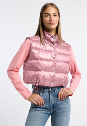 Jonge vrouw met een glanzend roze gewatteerde bodywarmer over een roze shirt met lange mouwen en een hooggetailleerde blauwe spijkerbroek, handen in de zij.