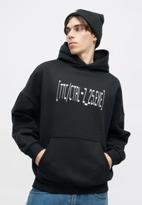 Μαύρη κουκούλα με μπροστινή τσέπη και λευκό εκτυπωμένο κείμενο που λέει «[TTC/CTRL-Z 2.5.EXE]». Συνδυασμένο με μαύρο καπέλο και μπλε τζην.