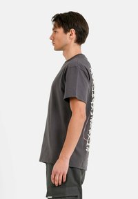 Jonge man in profiel, gekleed in een donkergrijze graphic T-shirt en donkere cargobroeken, tegen een eenvoudige witte achtergrond.