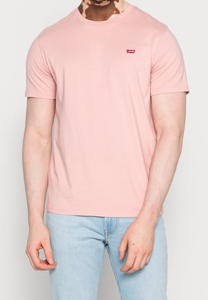 Person trägt ein hellrosa Levi's T-Shirt und hellblaue Jeans, steht mit entspannten Armen neben dem Körper vor einem einfarbigen Hintergrund.