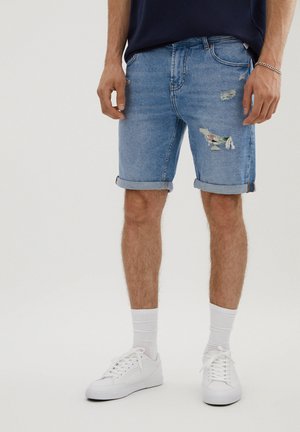 Homme portant un short en jean bleu clair retroussé avec des détails déchirés, des chaussettes blanches et des baskets blanches, debout devant un fond uni.