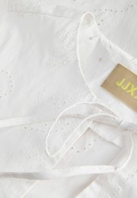 Vêtement blanc avec broderie ajourée, présentant un col rond, un détail noué et une étiquette beige avec du texte jaune.