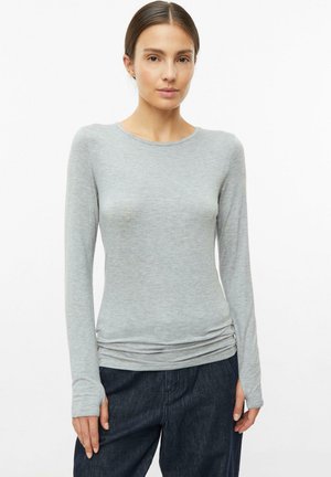 Young woman with dark hair wears a fitted light grey long-sleeve top and dark pants against a plain white background.