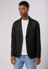 Černý blazer s klopami, předními nakládanými kapsami a zapínáním na knoflíky, vyrobený z texturované tkaniny. Nosený přes bílé tričko a džíny.