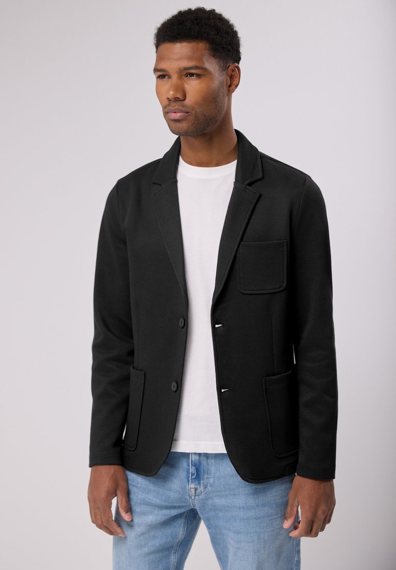 Černý blazer s klopami, předními nakládanými kapsami a zapínáním na knoflíky, vyrobený z texturované tkaniny. Nosený přes bílé tričko a džíny.