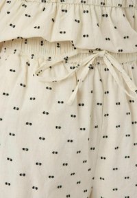 Tejido color crema con patrones de lazos negros, borde superior con volantes y una cintura ajustable con cordón para el ajuste del fit.