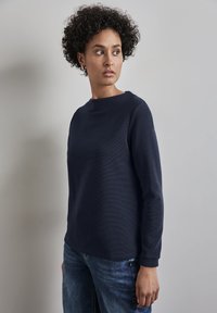Marinblå bomullsskjorta med lång ärm, rund halsringning, texturerat tyg och ribbade muddarna, parat med blå jeans.