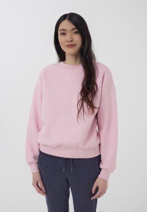 Giovane donna con lunghi capelli scuri, che indossa una felpa rosa e pantaloni della tuta grigio scuro, in piedi contro uno sfondo bianco uniforme.
