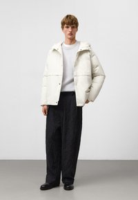 Vit isolerad jacka med huva, framknäppningar och sidofickor, bärs över en ljusgrå tröja och strukturerade svarta byxor.