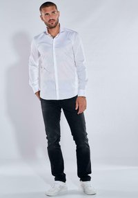 Weißes Langarmhemd mit Knopfleiste, kombiniert mit dunklen Jeans und weißen Turnschuhen, vor einem schlichten Hintergrund stehend.