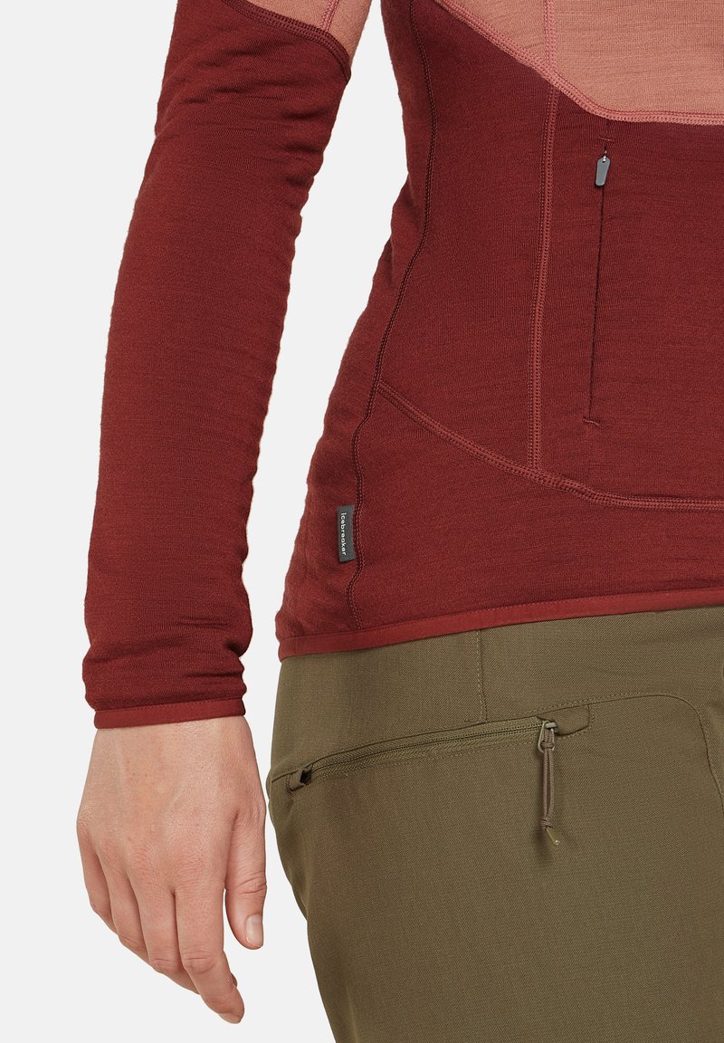 Langärmliges, geripptes bordeauxfarbenes Oberteil mit nahtlosem Design, ausgestattet mit einer Seitentasche und einem taillierten Schnitt, kombiniert mit grünen strukturierten Hosen.