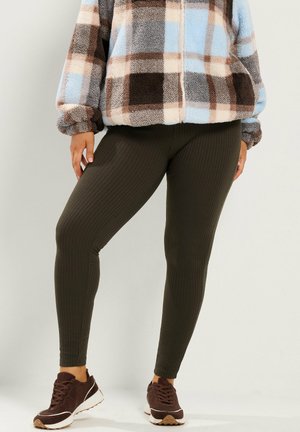 Personne portant des leggings sombres côtelés, des baskets marron et une veste polaire zippée à carreaux bleu, marron et beige.
