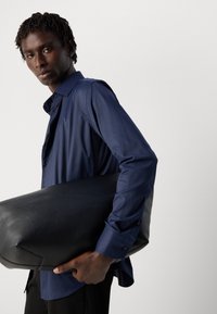 Camisa de manga larga azul marino con una textura suave, usada mientras sostiene un gran bolso de cuero negro suave. Diseño simple con acentos de botones.
