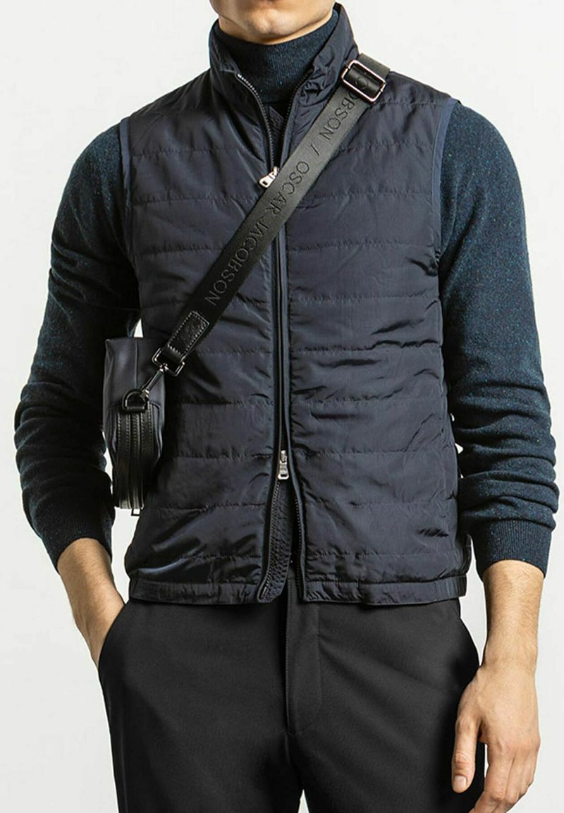 Marinblå quiltad väst med hög krage, framzipper och sidofickor. Bärs över en mörk tröja, tillsammans med svarta byxor och en liten väska.