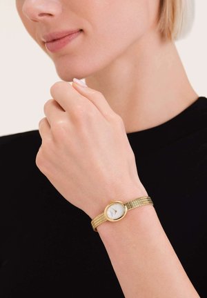 Gold-tone metal watch with a woven band, round white dial, and minimalist markings. Hand resting near the chin with a dark shirt background.