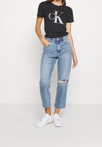 Czarny bawełniany t-shirt z błyszczącym logo "CK", zestawiony z jasnoniebieskimi jeansami o wysokim stanie z postrzępionymi detalami oraz białymi sneakersami.