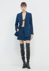 Femme portant un blazer en velours côtelé bleu et une jupe, un soutien-gorge en dentelle noire, et des bottes en cuir noires arrivant au genou, se tenant devant un fond blanc uni.